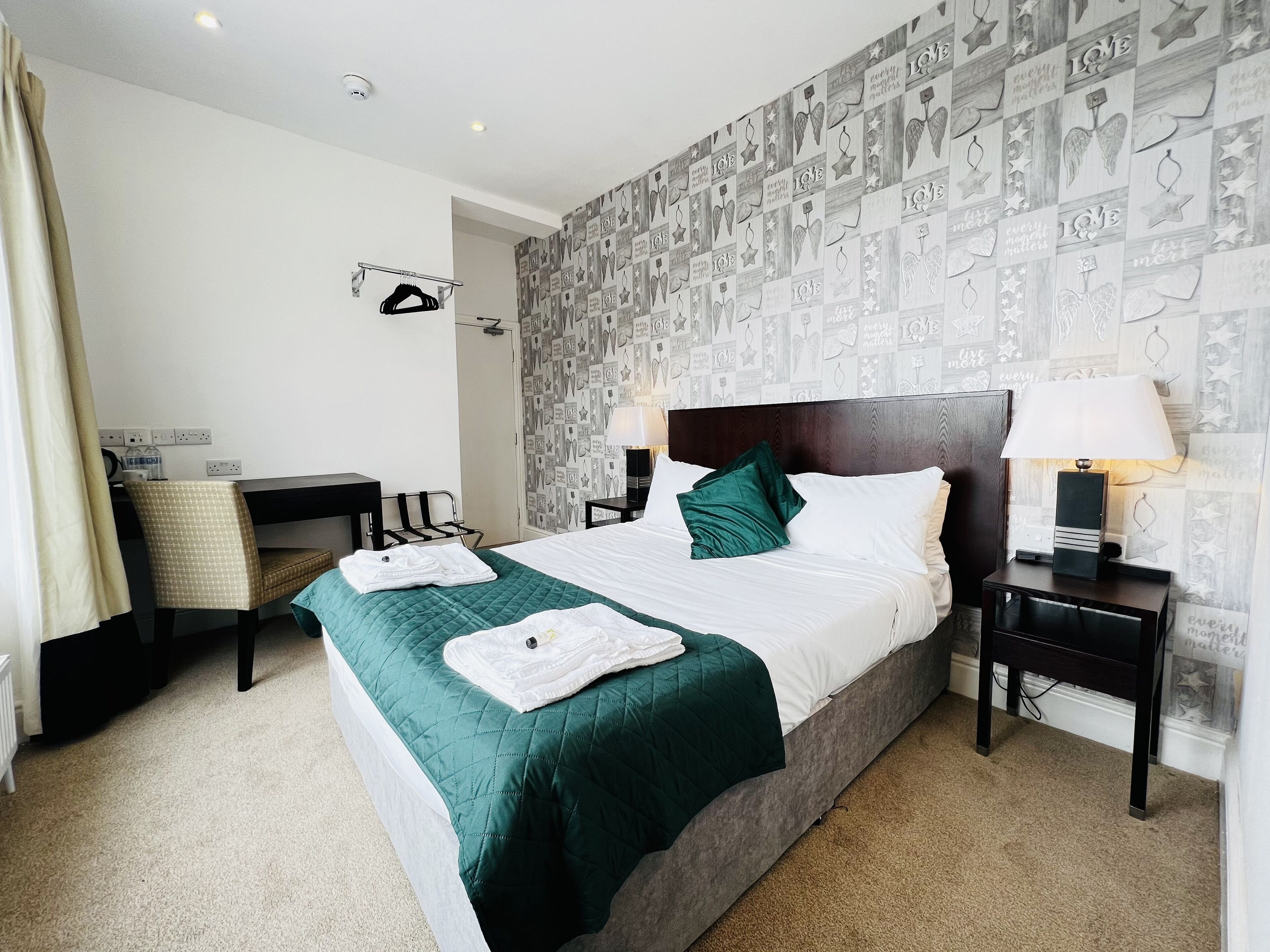 A hotel room with a large bed, two bedside tables with lamps, a desk with a chair, a wardrobe, and a patterned wallpaper.