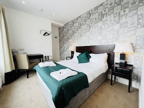 A hotel room with a large bed, two bedside tables with lamps, a desk with a chair, a wardrobe, and a patterned wallpaper. - The Springfield Fairbourne (Fairbourne)