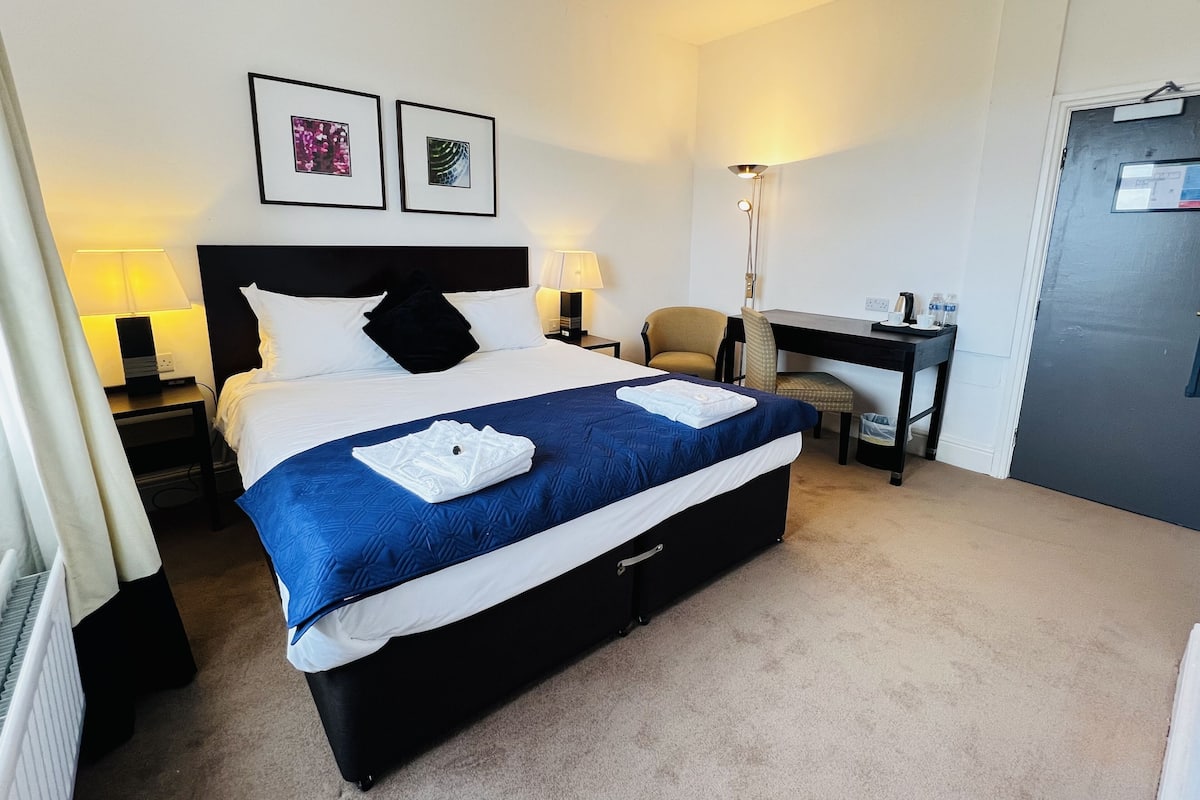 A hotel room with a bed, two bedside lamps, a desk, a chair, and two framed pictures on the wall.