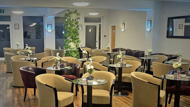 A dining area with multiple round tables and chairs, a large green plant, and wall-mounted lighting fixtures. - The Springfield Fairbourne (Fairbourne)