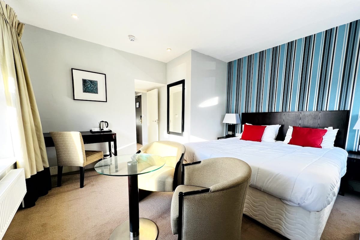 A hotel room with a large bed, a desk with a chair, a glass table, and a striped wall.