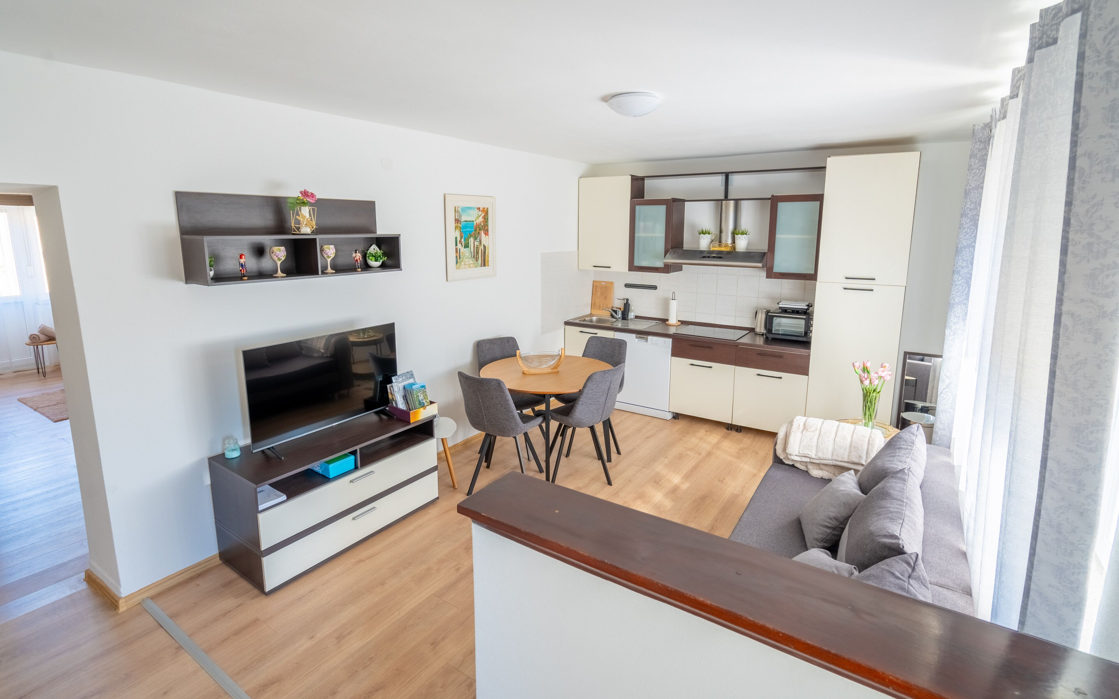 A modern living room with a flat-screen TV, a dining area with a round table and chairs, and a kitchen with white cabinets and a built-in oven.