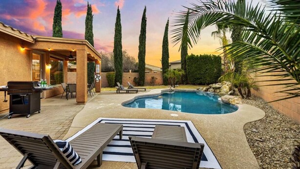 A backyard with a swimming pool, lounge chairs, a barbecue area, and tall trees.