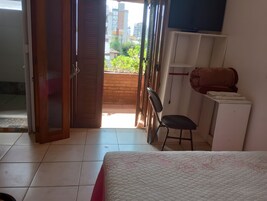Quarto de hotel com cama, escrivaninha, cadeira, televisão e varanda com vista para edifícios e vegetação.