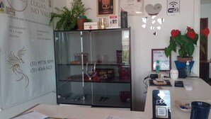 A reception desk with a glass partition, a potted plant, a framed certificate, and a QR code for check-in. - Merlyn Pousadas (Lajeado)