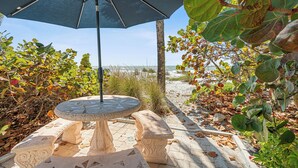 A stone table and bench under a blue umbrella, surrounded by foliage and a beach view. - Charming Beach Front Studio Steps to the Sand and Gulf (Longboat Key)