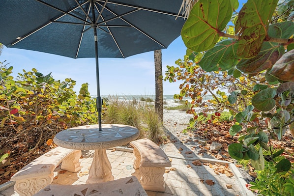 A stone table and bench under a blue umbrella, surrounded by foliage and a beach view.
