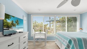 A bedroom with a TV on a dresser, a bed with a striped bedspread, a window with blinds, and a ceiling fan. - Charming Beach Front Studio Steps to the Sand and Gulf (Longboat Key)