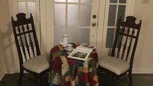 A dining area with two wooden chairs, a round table covered with a colorful tablecloth, a white teapot, and a magazine.