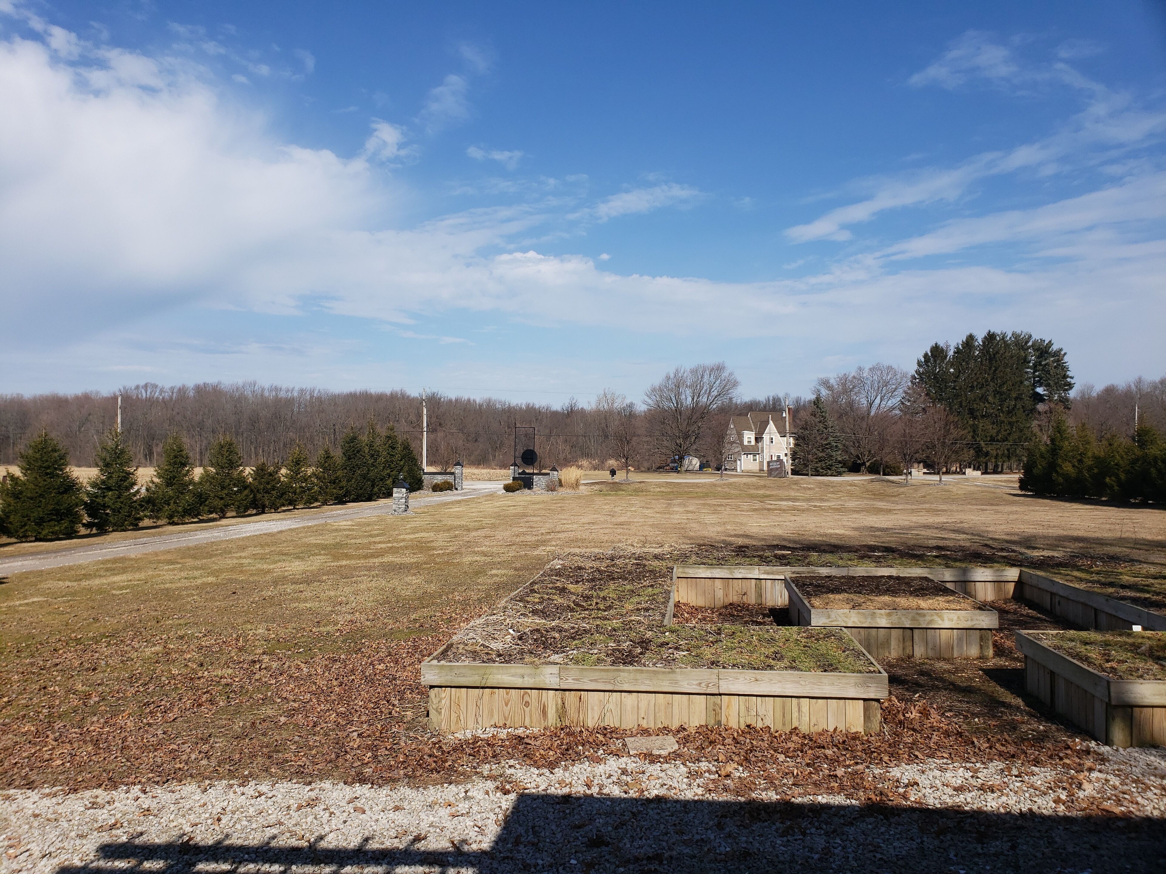A large open field with a wooden structure in the foreground, a white house in the background, and a forested area beyond.
