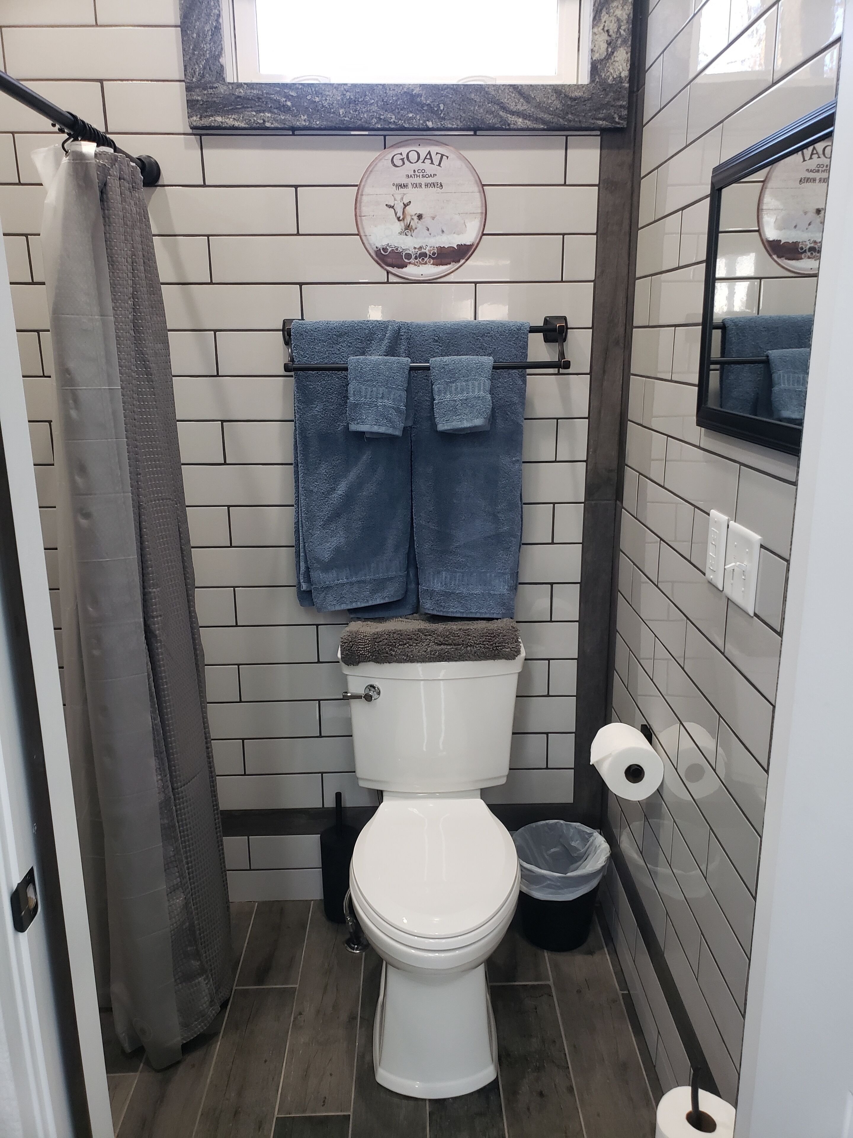 Un baño con inodoro blanco, toallero azul con dos toallas, dispensador de papel higiénico y un cubo de basura.