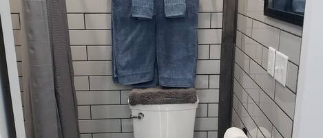 Un baño con inodoro blanco, toallero azul con dos toallas, dispensador de papel higiénico y un cubo de basura.