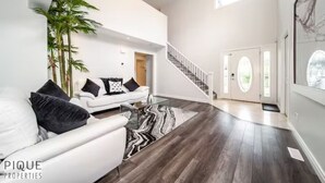 A spacious living room with a white sofa, a glass coffee table, and a large plant. A staircase leads to a second floor, and a door with a glass panel is visible. - 6B Luxury Home - Patio✔AC✔King Bed☆Park - WiFi (Edmonton)