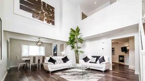 A spacious living area with a high ceiling, a dining table with chairs, two white sofas with black cushions, a large plant, and a fireplace. - 6B Luxury Home - Patio✔AC✔King Bed☆Park - WiFi (Edmonton)