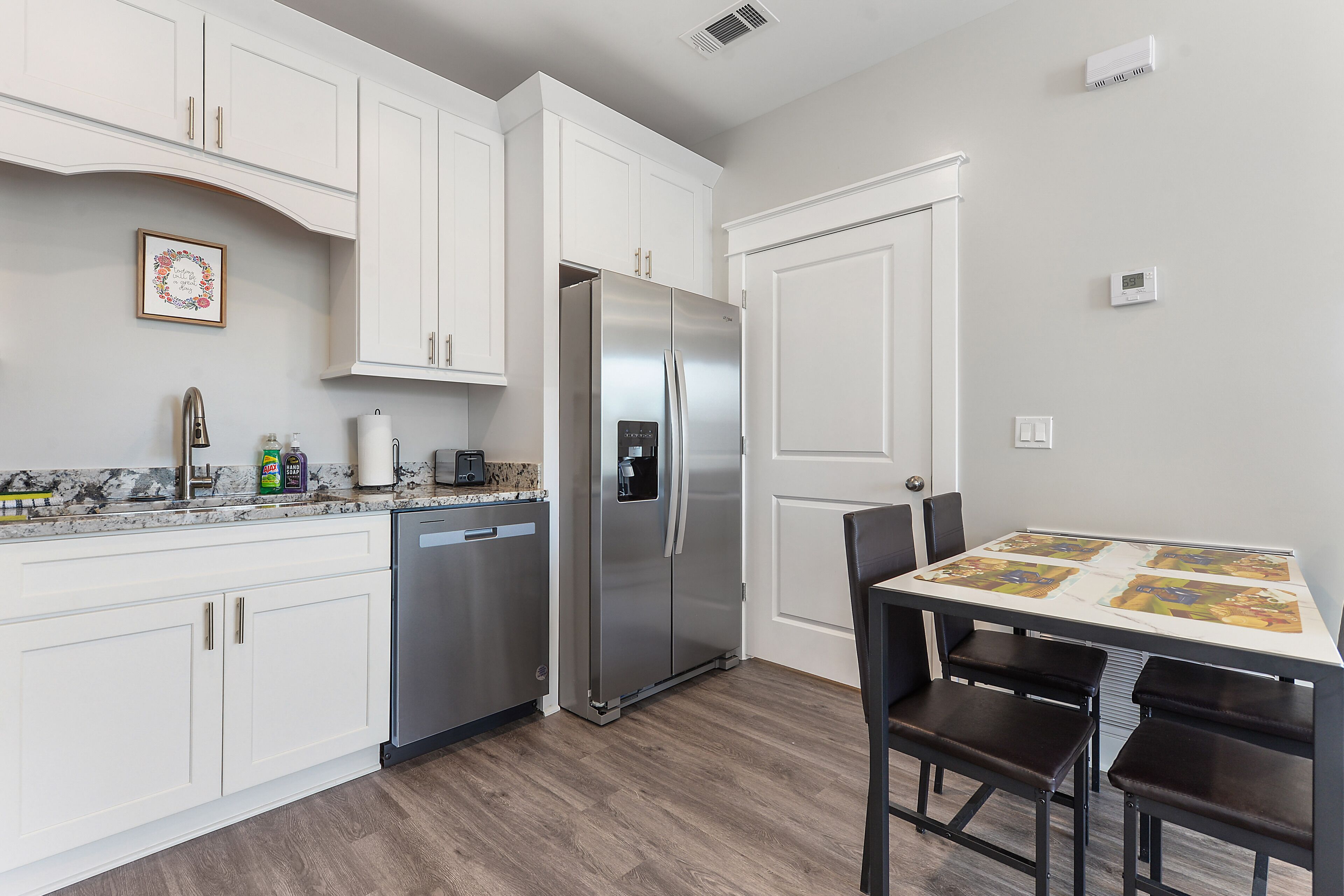 A modern kitchen with white cabinets, a stainless steel refrigerator, a dishwasher, and a dining table with four chairs.