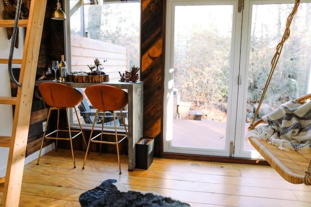 A cozy room with a wooden floor, a dining area with two chairs, a hanging swing, and a large window with a view of trees.