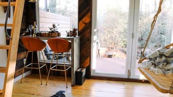 A cozy room with a wooden floor, a dining area with two chairs, a hanging swing, and a large window with a view of trees.