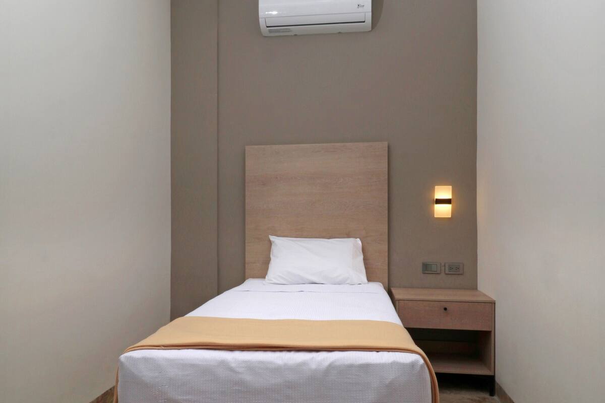 A hotel room with a bed, a nightstand, an air conditioning unit, and a wall-mounted light.