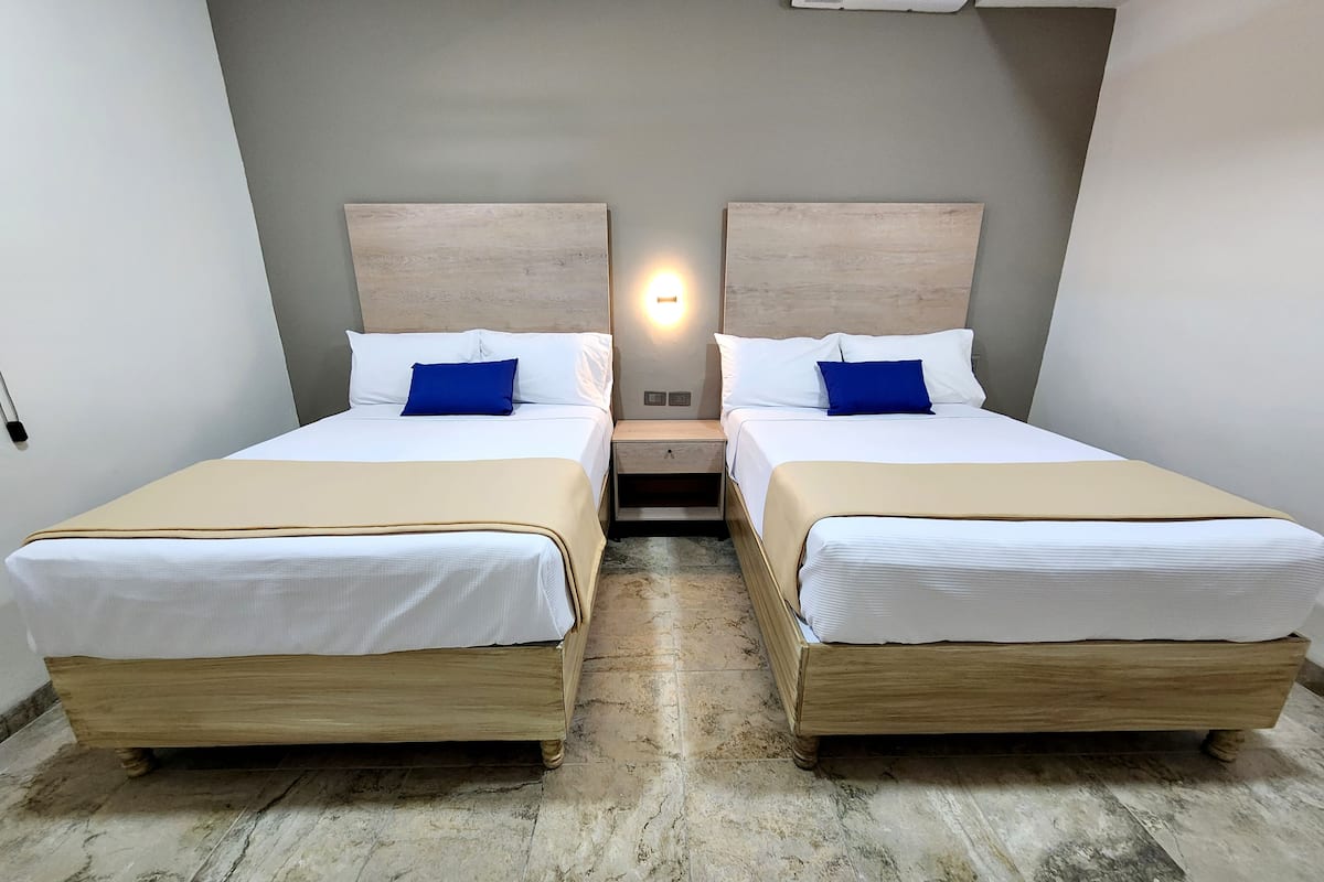 A hotel room with two beds, each with white linens and blue pillows, wooden headboards, and a nightstand between them.