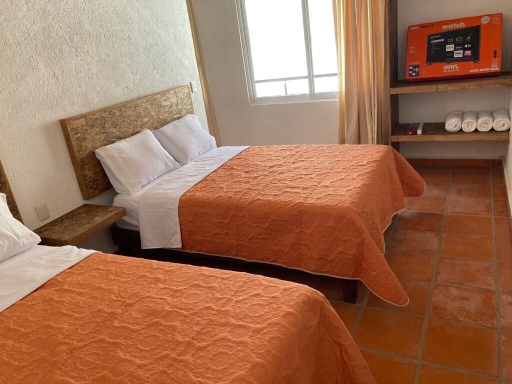 Two beds with orange bedding, a wooden headboard, a TV on a shelf, and a window with curtains.