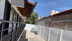 Un étroit couloir extérieur doté d’une rambarde en métal, une voiture blanche garée au bout, et un mur blanc à la surface texturée.