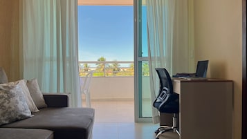 Una habitación con sofá, escritorio con computadora portátil y vista a palmeras desde un balcón.