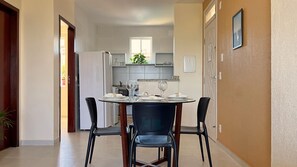 A dining area with a round table set for four, two black chairs, a white refrigerator, and a kitchen in the background. - 2 suítes ideal p/ amigos e casais (Aquiraz)