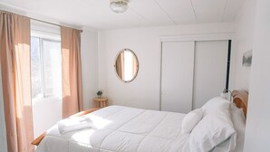 Un llit ben fet en una habitació amb finestra, un mirall rodó petit i una porta.