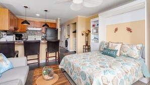 A modern kitchen with wooden cabinets, a dining area with a table and chairs, a bedroom with a bed and decorative pillows, and a ceiling fan.