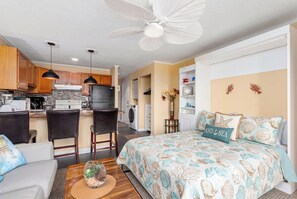 Cozinha moderna com armários de madeira, área de jantar com mesa e cadeiras, quarto com cama e almofadas decorativas, e um ventilador de teto.