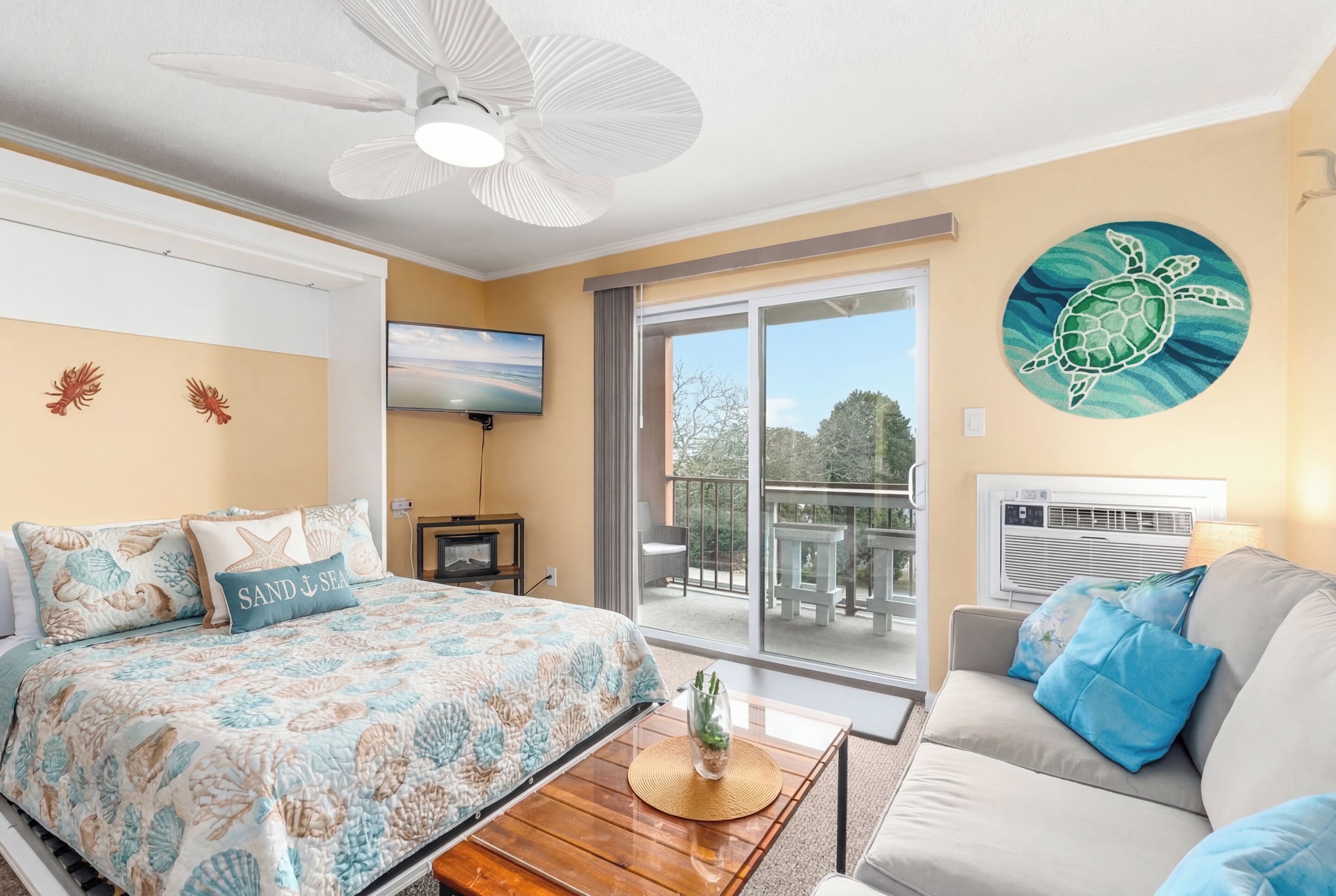 A bedroom with a bed, a ceiling fan, a TV, a balcony with a view, and a wall painting of a turtle.