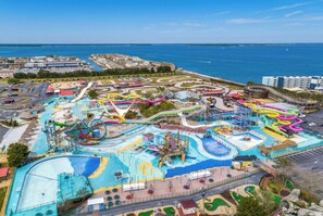 Vista aérea de un parque acuático con diversas toboganes y piscinas, rodeado de edificios y una masa de agua.