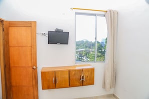 A room with a wooden door, a television mounted on the wall, a window with a curtain, and a wooden cabinet below the window.
