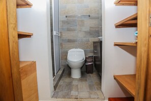 A narrow bathroom with a toilet, a trash bin, and a shower area.