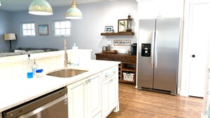 A kitchen with a dishwasher, sink, and a refrigerator. There is a window with blinds, a shelf with decorative items, and a hanging light fixture. - Blue Bayou - Charming home with heated pool, tiki and OPTIONAL BOAT DOCKAGE (Bradenton)