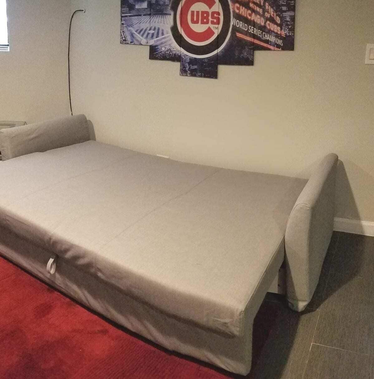 A single bed with a grey mattress and pillows, a red rug, and a wall with a Cubs-themed poster.