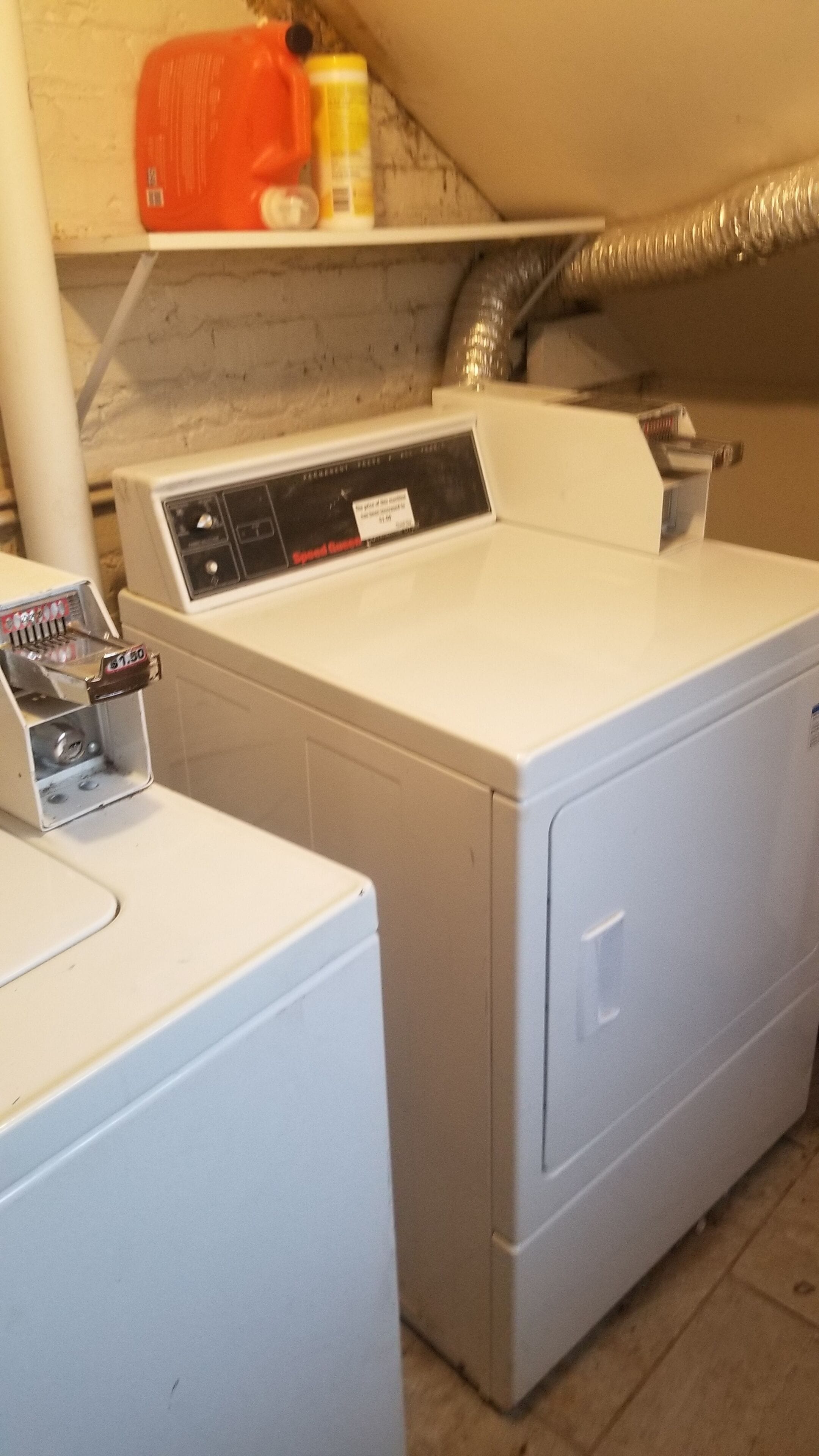A laundry area with two washing machines, a shelf with cleaning supplies, and a vent pipe.