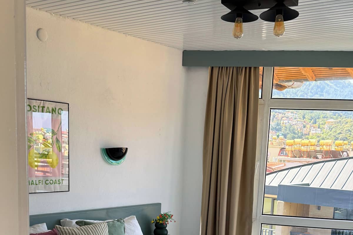 Habitación de hotel con cama, mesita de noche, lámpara de pared, cortinas y vistas a edificios.