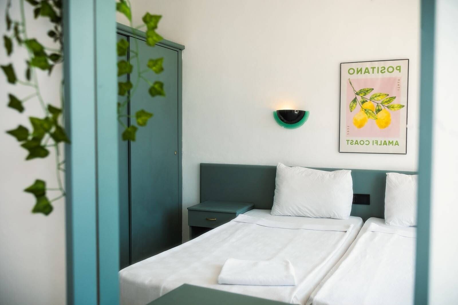 Quarto de hotel com cama, mesa de cabeceira, guarda-roupa verde e uma luminária fixa na parede.