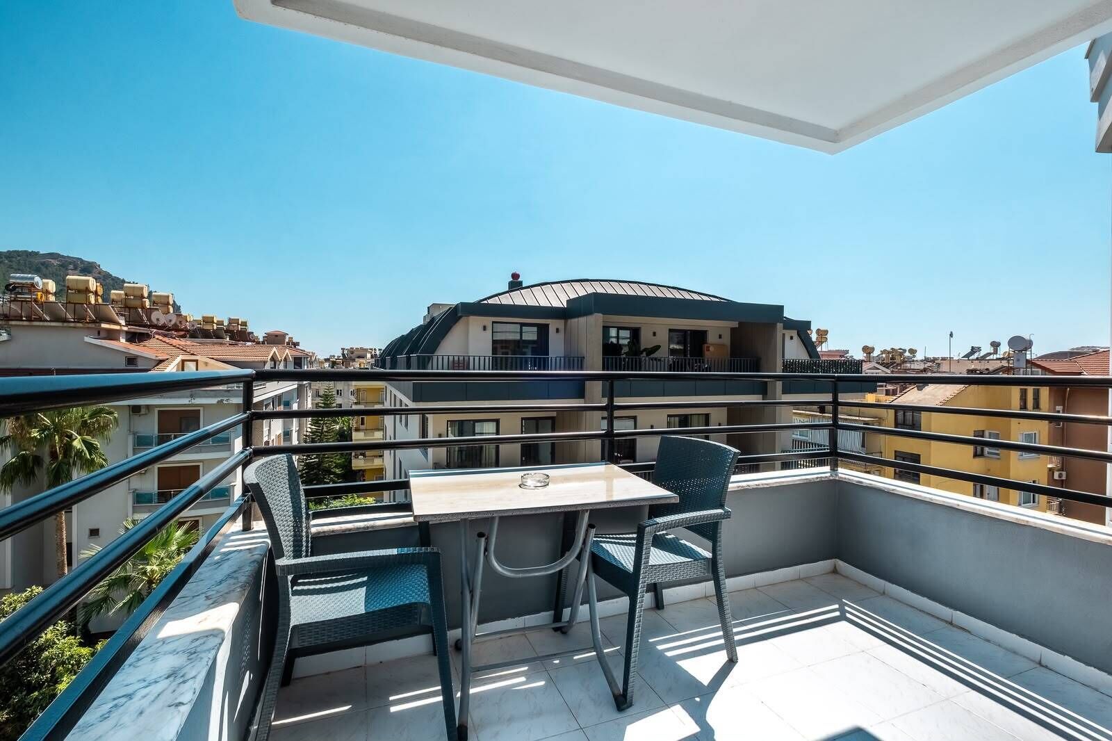 Un balcon avec une table et des chaises, donnant sur un paysage urbain avec des bâtiments et un ciel dégagé.