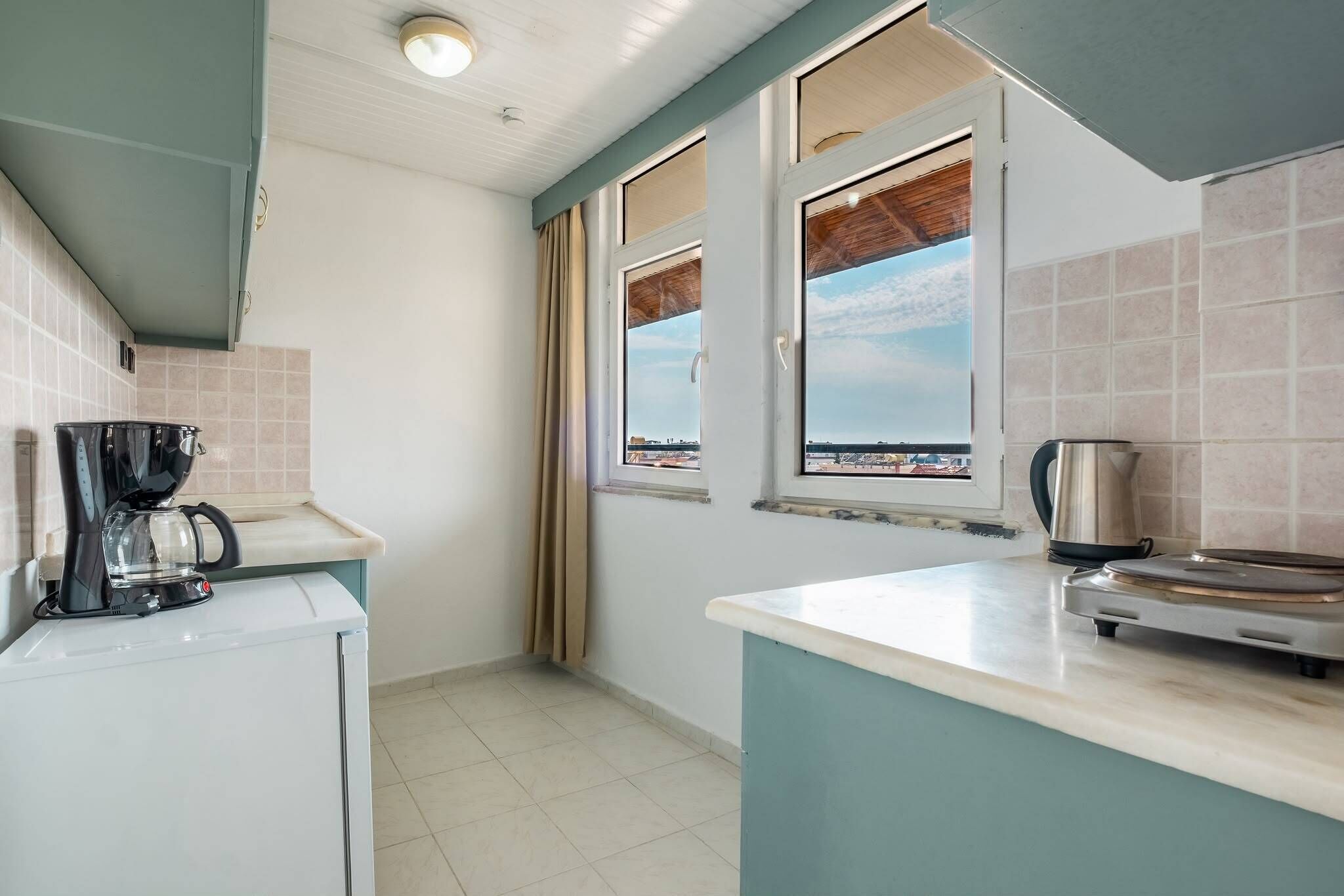 A compact kitchen with a coffee maker, a kettle, and a stove. There are two windows with curtains, and the kitchen has a tiled backsplash.