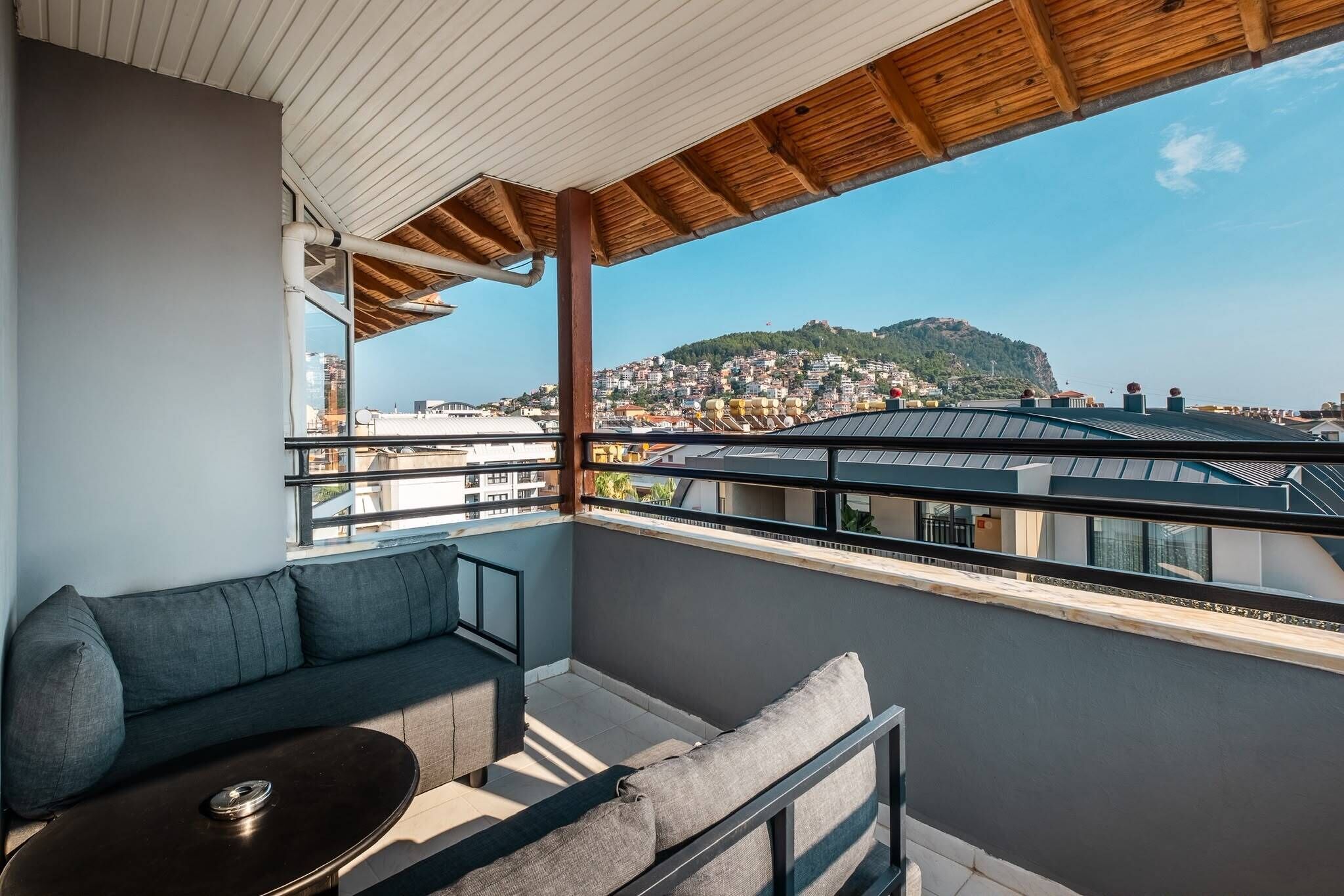 Ein Balkon mit einer Sitzecke und einem Tisch, mit Blick auf eine Stadtlandschaft mit Gebäuden und einer Anhöhe.