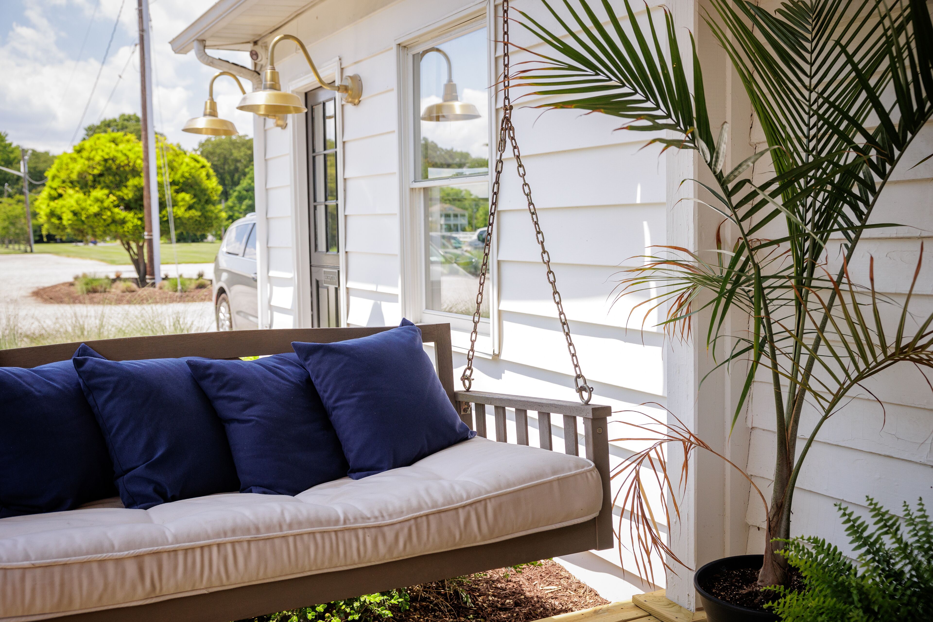 A porch with a swing seat, a potted plant, and a view of a parked car and trees.