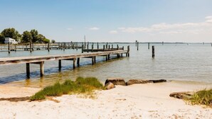 Een houten steiger die zich uitstrekt over rustig water, een zandstrand met schaarse begroeiing en een heldere lucht.