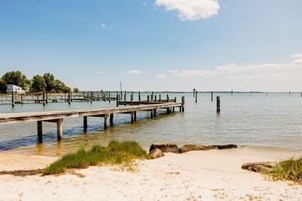 A wooden pier extending into calm waters, a sandy beach with sparse vegetation, and a clear sky.