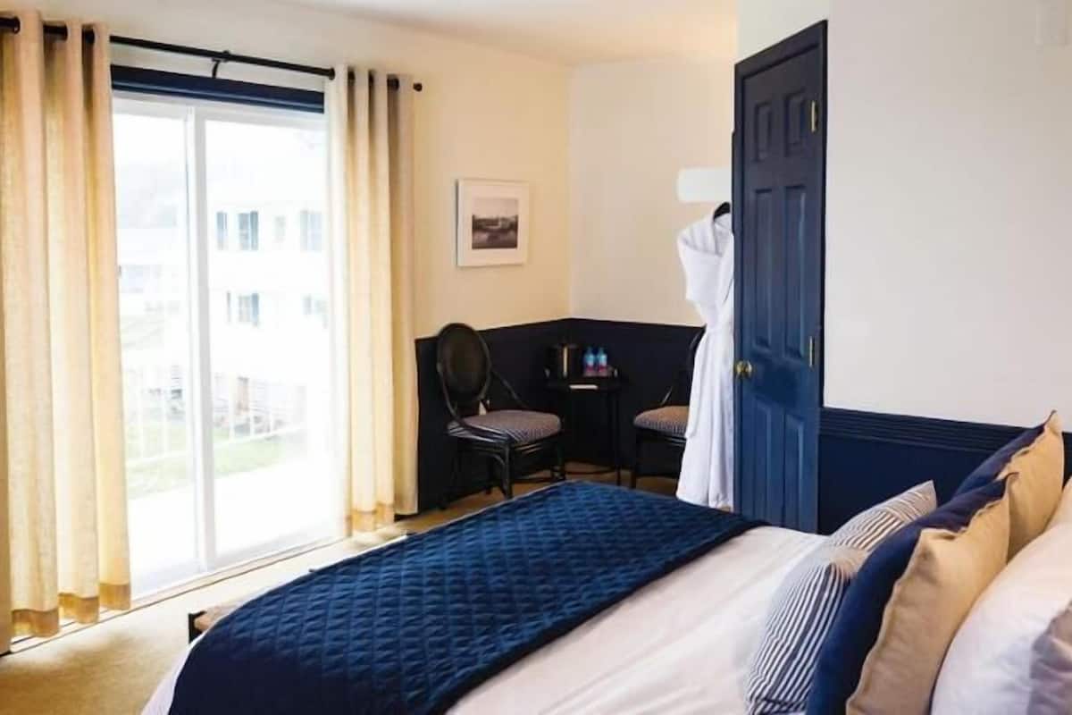 Une chambre d’hôtel avec un lit, un bureau et une chaise. Il y a une fenêtre avec des rideaux, une porte et un tableau au mur.
