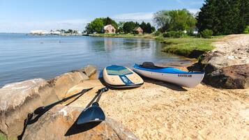 Un kayak et une pagaie sur une plage de sable, au bord d’une étendue d’eau calme, avec des maisons en arrière-plan.