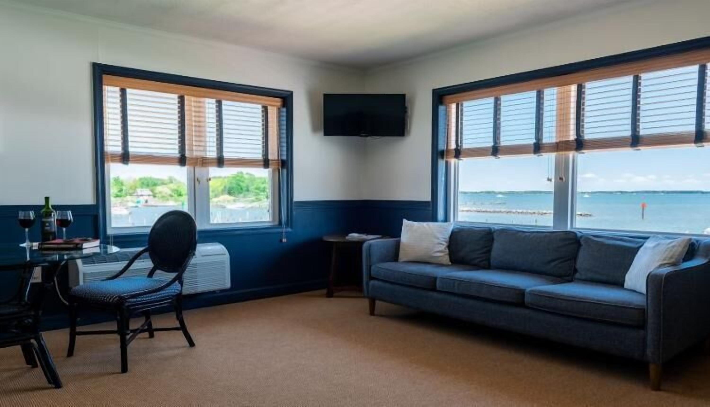 A room with a sofa, a chair, a small table, and a TV. There are windows with blinds and a view of the sea.