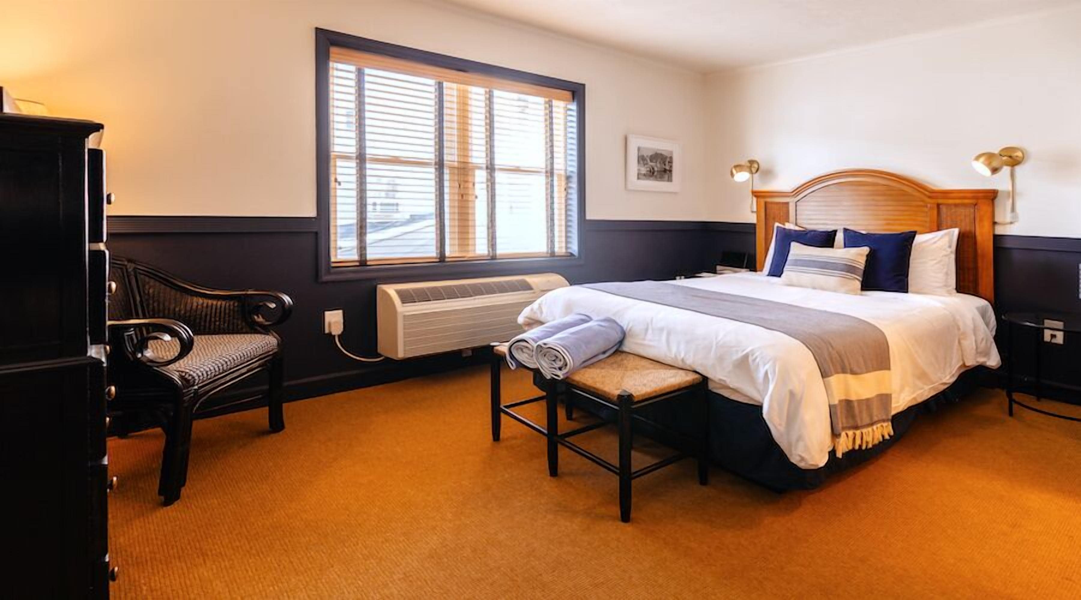 A hotel room with a large bed, a bench, a desk, and a window with blinds.