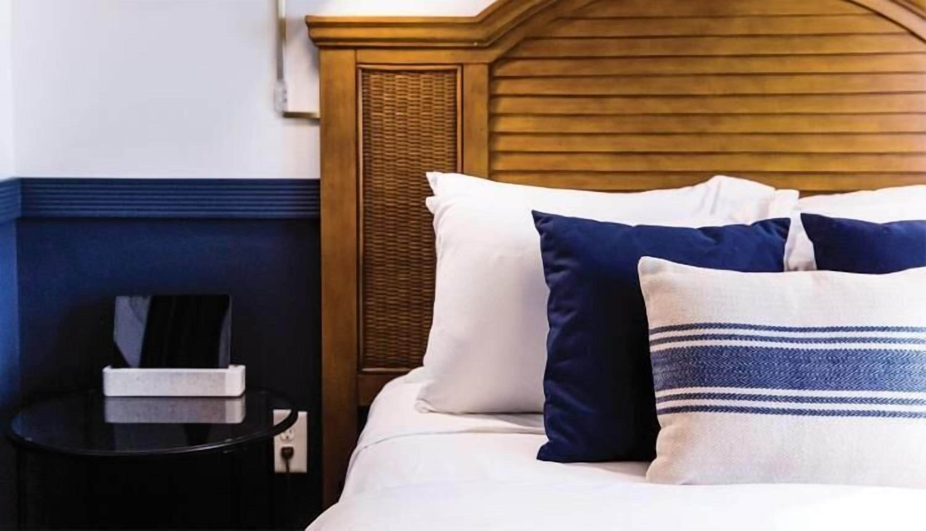 Una cama bien tendida con almohadas blancas y azul marino, un cabecero de madera y una mesita de noche con un libro y una caja.